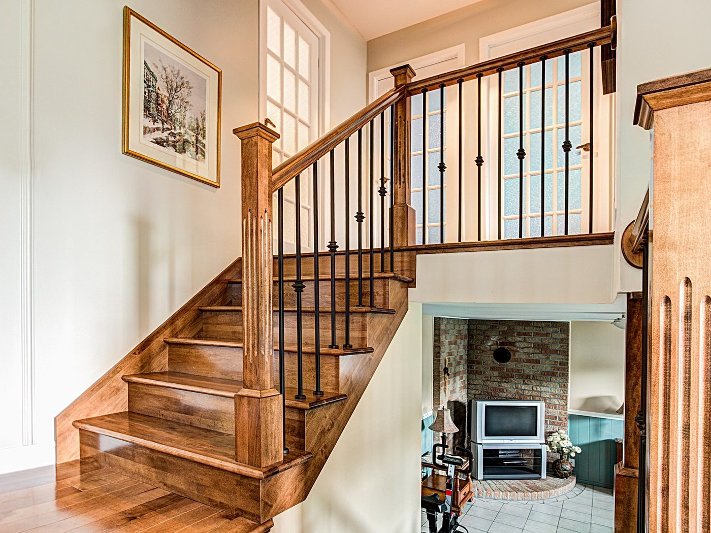 Un escalier de chêne à poteaux carrés et barreaux de fer forgé, tableaux au mur, salon en contrebas