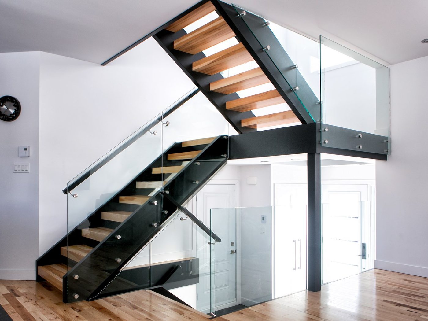 Un escalier à limon d'acier noir et marches de bois, ouvert, garde-corps de verre, horloge au mur