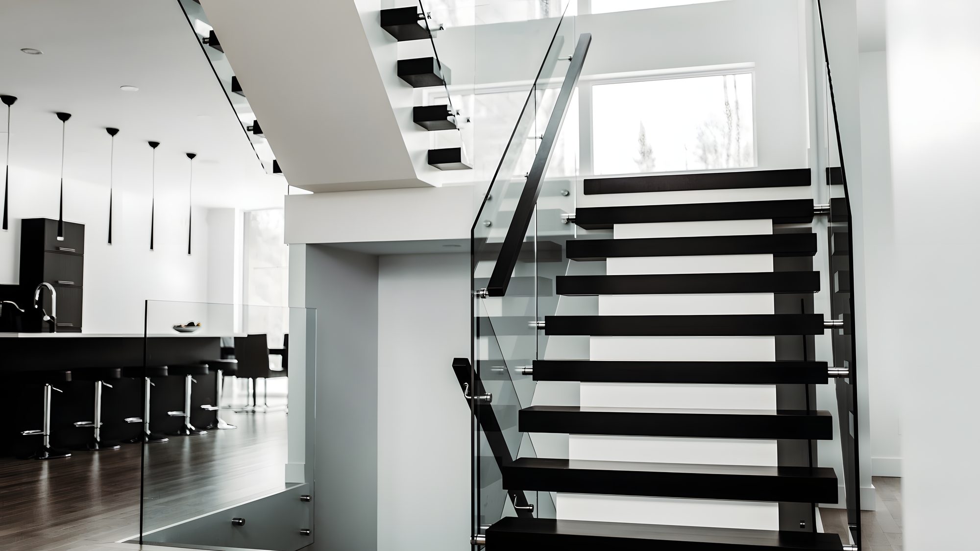 Un escalier aux marches noires suspendues entre deux panneaux de verre, dans une maison aux murs blancs, la cuisine sombre à gauche