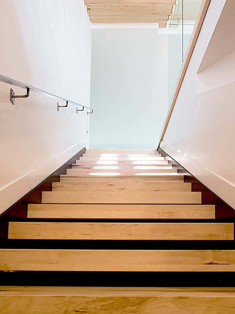 Un escalier d'érable clair aux marches ouvertes, vu d'en bas, main courante d'acier sur mur blanc