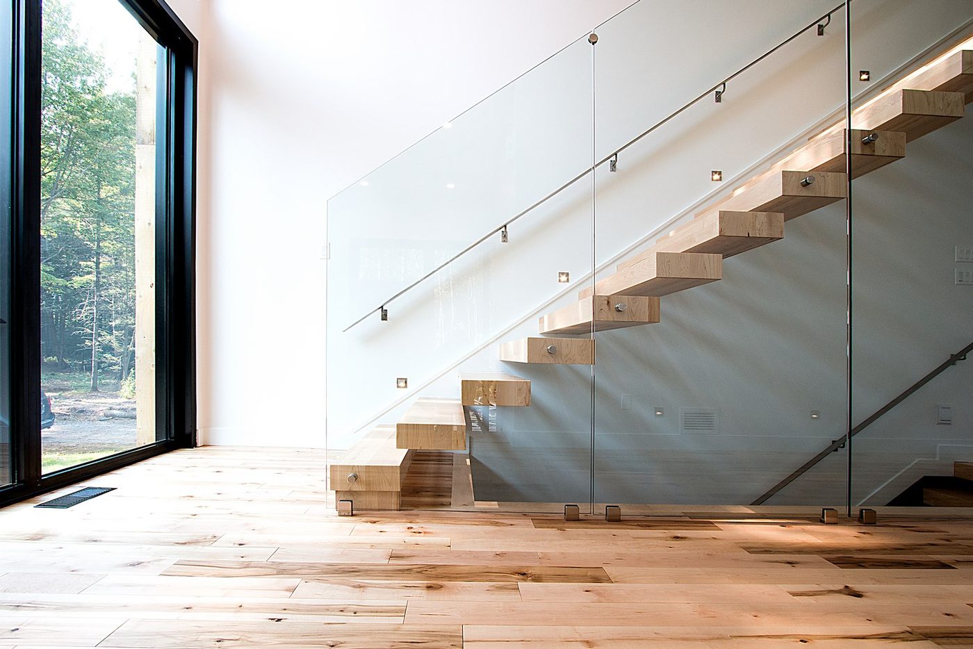 Un escalier d'érable clair à garde-corps de verre devant de grandes fenêtres noires, forêt derrière