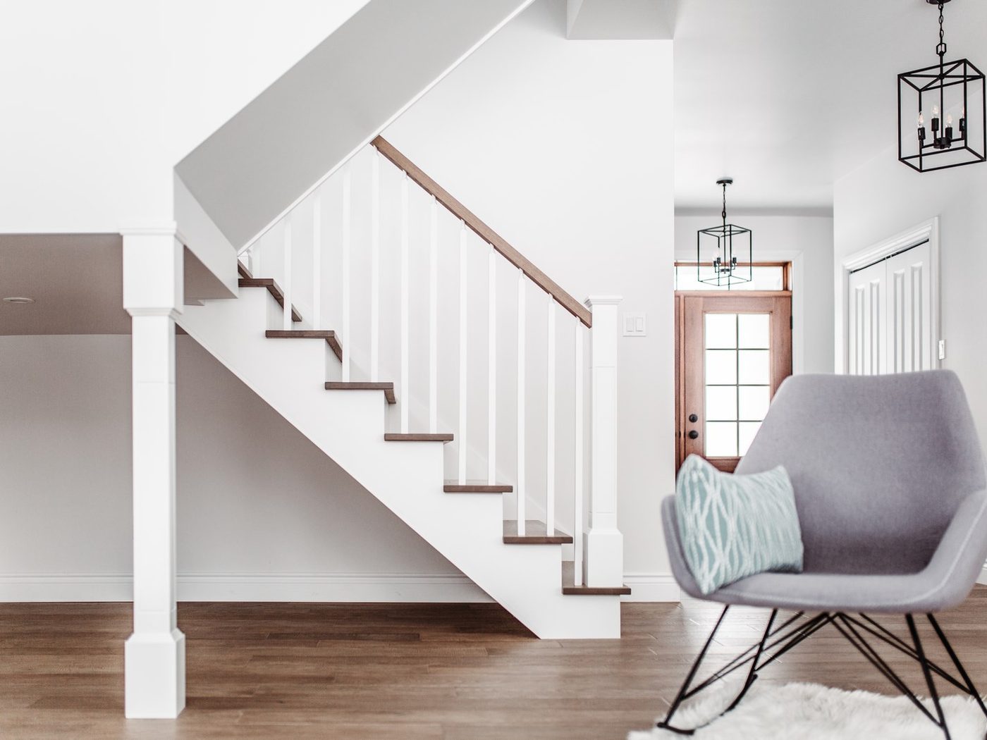 Un escalier blanc à main courante de bois dans un salon, fauteuil gris et tapis blanc au pied